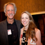 A man and woman smiling at the camera, both wearing name tags. The man is in a dark blazer and black shirt, while the woman is in a light jacket over a patterned top. They are standing in a warmly lit room with brown textured walls.