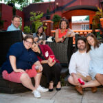 A group of people sitting and kneeling on a patio, some holding drinks and smiling at the camera. They are casually dressed and appear to be enjoying a social gathering with a mix of outdoor and indoor seating.