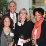 Four people are standing together, smiling at a social event. They are dressed in business attire, holding wine glasses. A large indoor space with trees and people is visible in the background.