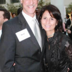 A man and a woman, both wearing name tags, pose closely for a photo in a formal setting. The man is in a dark suit with a tie, and the woman is in a black outfit. They're smiling with people visible in the blurred background.