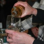 A person pouring white wine from a bottle into a glass. The setting appears to be a tasting event with other bottles in the background. The person is wearing a black jacket and a light-colored patterned garment.