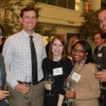 A group of five people standing together at an indoor event, holding wine glasses and smiling at the camera. They are dressed in business attire, and name tags are visible on some of their chests.