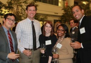 A group of five people standing together at an indoor event, holding wine glasses and smiling at the camera. They are dressed in business attire, and name tags are visible on some of their chests.