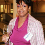 A person wearing a textured jacket and purple top holds an engraved wine glass. They have a name tag on their jacket. The background shows an indoor setting with trees and a lit hallway.