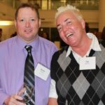 Two people at an indoor event, both wearing name tags. The person on the left is dressed in a purple shirt and striped tie, holding a glass. The person on the right has short, light-colored hair and wears a diamond-patterned sweater over a collared shirt.