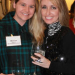 Two women smiling and holding wine glasses at an indoor event. The woman on the left is wearing a green checkered shirt with a name tag. The woman on the right is wearing a black top with a patterned turtleneck.