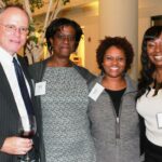 Four people stand together indoors, smiling. The person on the left holds a glass of wine. All are wearing name badges. They are dressed in business or semi-formal attire. A decorative indoor plant is in the background.