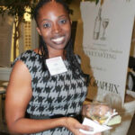 A woman smiling at a wine tasting event, holding a glass of white wine. She wears a patterned dress and has a name tag. A sign in the background reads "Wine Tasting.