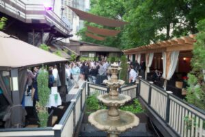 Outdoor event with a crowd of people gathered on a patio surrounded by trees and buildings. A tiered fountain is in the foreground, and tented spaces with people socializing can be seen along the sides.