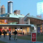 A modern building with a curved facade and glass tower stands illuminated at dusk. People walk toward the entrance, with skyscrapers visible in the background. A sign promoting Coke is seen near the walkway.