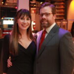 A smiling couple poses for a photo at an indoor event. The woman wears a black dress, and the man is in a gray suit with a red tie. The background shows other people and a warmly lit venue.
