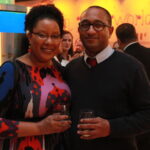 A woman and a man stand closely together at a social event, smiling and holding drinks. The setting is a colorful room with blurred figures and decorations in the background.