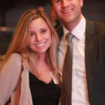 A man and woman smiling, standing close together. The woman is wearing a black top with a light jacket and holding a phone. The man is in a dark suit with a tie. They appear to be at an evening event outdoors.