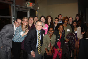 A diverse group of people, dressed in business attire, pose together indoors at a social event or gathering. They are smiling and standing closely for the photo.