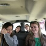 Four women are sitting in the back of a van. Three are smiling at the camera, and one is playfully covering her face with her hand. The interior is brightly lit, and trees and a building are visible through the windows.