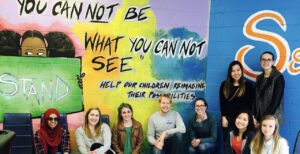 A group of people is sitting and kneeling in front of a colorful mural on a wall. The mural includes the phrases 