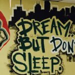 A mural featuring a hand gesture on the left and bold graffiti text reading "Dream... But Don't Sleep" against a city skyline backdrop. The right side includes additional small text and colorful paint splashes.