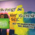 Colorful mural with the text "You can not be what you can not see." A child holds a sign that says "Stand." Additional text reads "Help our children reimagine their possibilities." Bright and motivational artwork. Blue chairs are in front of the mural.