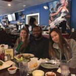 Three people are sitting together at a restaurant table with plates of food and drinks in front of them. The background features a vibrant mural and a bar area with bottles displayed on shelves.