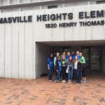 A group of people stand smiling in front of the entrance to Thomasville Heights Elementary school. They are casually dressed and appear to be a mix of adults and teenagers. The building's address, 1820 Henry Thomas, is visible above the entrance.