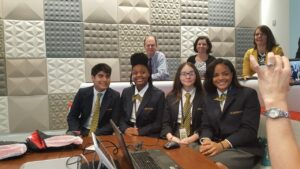 A group of four students in matching uniforms sit at a table with laptops, smiling for a photo. Behind them, three adults are seated, also smiling, in a room with geometric patterned walls. A person in the foreground is capturing the scene on a phone.