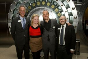 Four people stand smiling in front of a large vault door. The two men on the ends wear suits, the woman wears a red and tan dress, and the man next to her wears a gray suit. They appear to be at a formal or professional event.