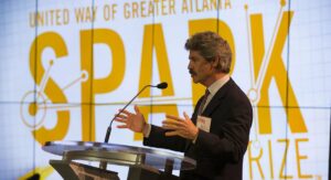 A man with a mustache speaks at a podium in front of a large 