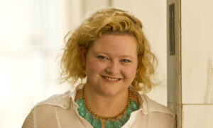 A person with curly blonde hair smiles while leaning against a light-colored wall. They are wearing a white blouse and a necklace with large turquoise beads. The background is softly lit.