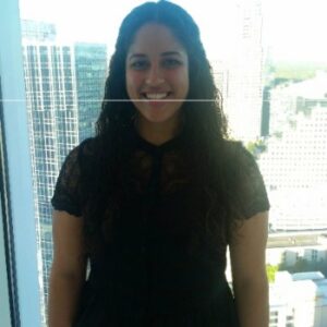 A person with long, dark hair stands smiling in front of a window in a high-rise building. The cityscape with modern buildings is visible in the background. The person is wearing a black lace top.