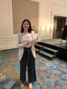 A woman stands smiling in a room with patterned carpet and a stage in the background. She is wearing a polka dot outfit with a light cardigan and is holding a glass award.