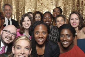 A group of people smiling and posing together in front of a gold sequin backdrop. They are dressed in formal and semi-formal attire, with expressions of joy and camaraderie.