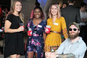 Four people smiling at a social gathering. Three women stand, holding drinks, with name tags on their outfits. A seated man with a beard and tattoos wears sunglasses and a cap. People are chatting in the blurred background.