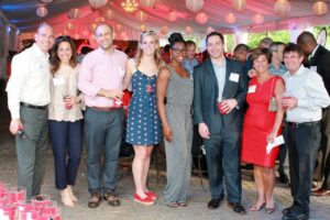 A group of eight people, dressed in semi-formal attire, are standing together and smiling at an event under a tent. Some are holding red plastic cups. The ceiling is decorated with paper lanterns and drapes.