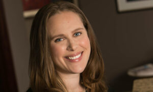 A person with long, light brown hair smiles at the camera. They are in a dimly lit room with blurred background elements. They are wearing a dark top and have a cheerful expression.