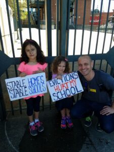 Two children holding signs about stable homes and evictions, standing with a smiling man. They are in front of a metal gate outside, with buildings visible in the background.