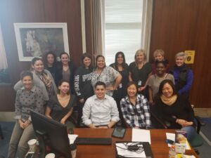 A group of sixteen people posing together in an office setting. They are smiling and seated or standing around a desk with a computer and papers. The room has wooden walls and a large window with blinds.
