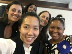 A group of five people smiling and taking a selfie indoors. They are standing close together and appear to be in a casual setting, possibly an office or conference room.