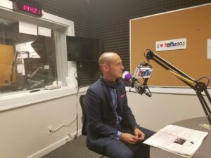 A person is seated in a radio studio, speaking into a microphone. They are wearing a suit and have headphones around their neck. The studio walls are covered in soundproofing foam, and there is a window to another room.