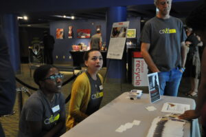 Two people sitting at a table with brochures and a poster, wearing matching t-shirts. A man stands nearby. The background shows a recycle bin and art displays. The setting appears to be an event or exhibition.