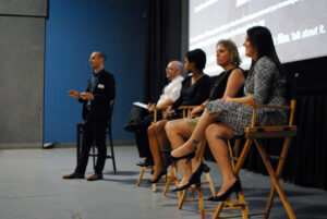 A panel discussion with five people is taking place. One person is standing and speaking, while four others are seated on chairs, listening intently. A large screen is visible in the background.