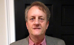 A middle-aged man with light hair is wearing a gray suit jacket and a red shirt. He stands in front of a dark door, looking directly at the camera.