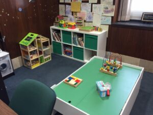 A playroom with a green tabletop featuring colorful toys, including a bead maze and building blocks. There's a storage unit with books and bins against a wooden wall decorated with children's drawings. A small toy kitchen set is in the corner.