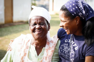 Two women smiling warmly at each other. The older woman, wearing a white headscarf and apron, has her eyes closed. The younger woman, in a patterned bandana and T-shirt, is embracing her. They appear to be outside on a sunny day.