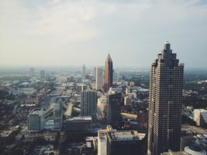 Aerial view of a cityscape with several tall buildings and skyscrapers under a clear sky. The foreground shows a mix of modern architecture, roads, and green spaces, with a central, prominent, triangular skyscraper standing out in the middle distance.