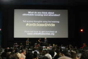 A panel discussion in a dimly lit theater. The screen behind shows a prompt about affordable housing and education, encouraging audience participation via a hashtag. People are seated, listening attentively.