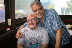 Two smiling men pose together indoors. One man is seated wearing a t-shirt with a colorful design, while the other stands beside him in a checkered shirt, resting his arm on the seated man's shoulder. They appear relaxed and happy in a casual setting.