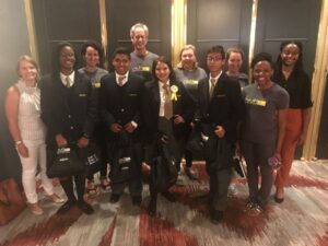 A group of people standing together in a room. Some are wearing matching jackets, shirts, and ties, while others are in casual attire with T-shirts. They are smiling and holding bags. The background features doors with gold trim.
