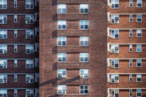 Close-up view of a brick apartment building with multiple identical windows. Air conditioning units are visible beneath several windows. Shadows cast by a nearby structure create geometric patterns on the building facade.