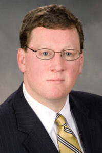 A man wearing glasses and a dark suit with a yellow tie is looking at the camera. The background is a neutral gray.