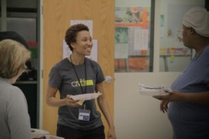 A smiling person wearing a gray t-shirt holds a plate of food, engaging with another person in a blue shirt who is also holding a plate. They are in a room with educational posters on the walls.